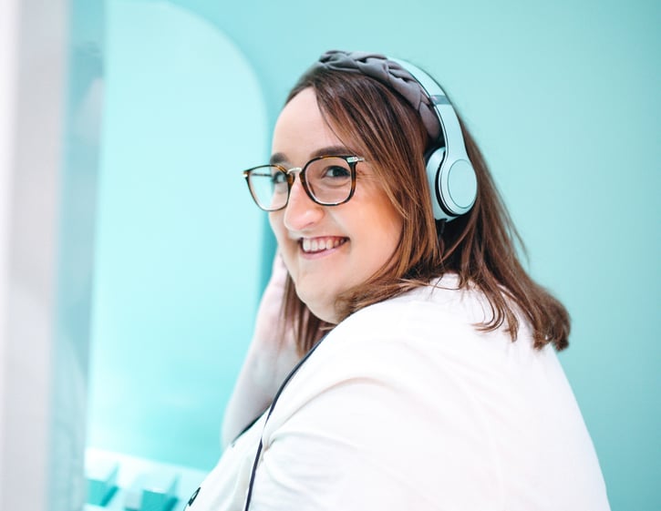 Woman wearing blue headphones and a white tshirt in front of a blue wall. She is smiling at the camera.