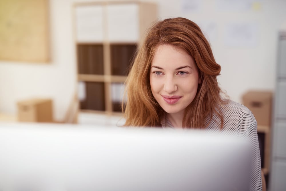 Attractive young woman working on a desktop computer smiling as she leans forwards reading text on the screen, view over the monitor-3