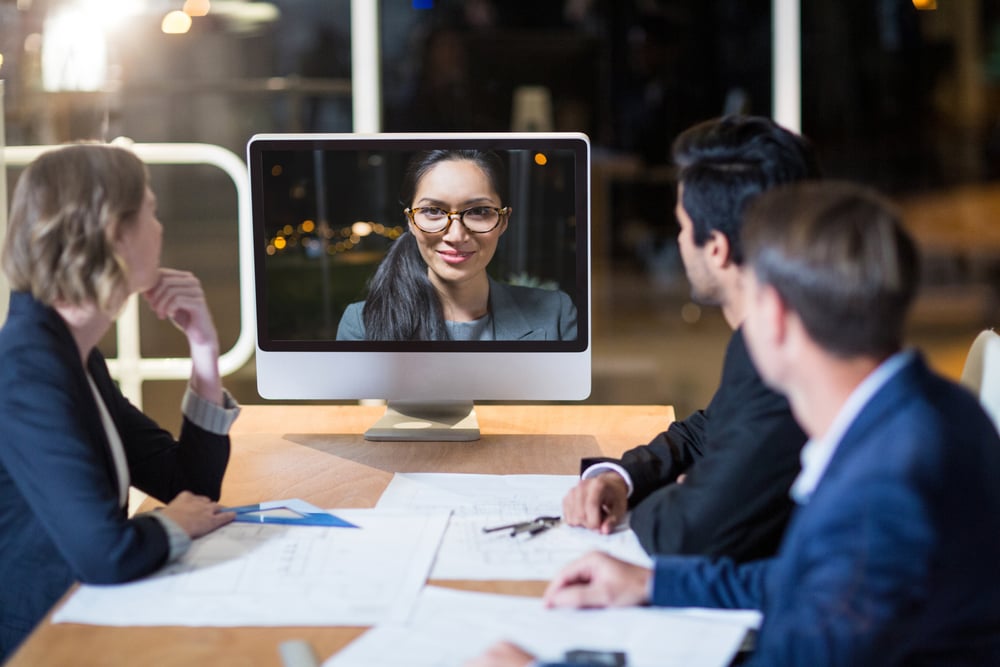 in Team von Mitarbeitern arbeitet im modernen Büro, während ein Kollege über Videokonferenz aus dem Homeoffice zugeschaltet ist 