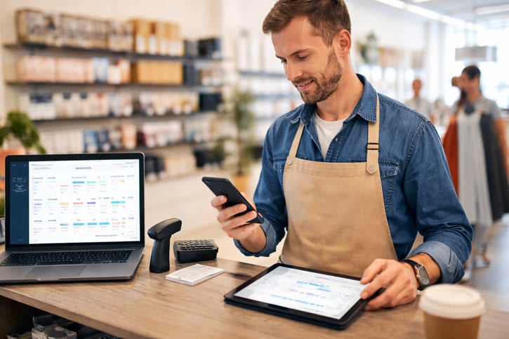 Store manager approving staff holiday requests on a mobile app while checking the rota on a tablet behind the shop floor counter.