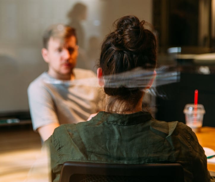 Man en vrouw zijn in gesprek. We kijken over de schouder van de vrouw mee en zien haar gesprekspartner, die niet in focus is.