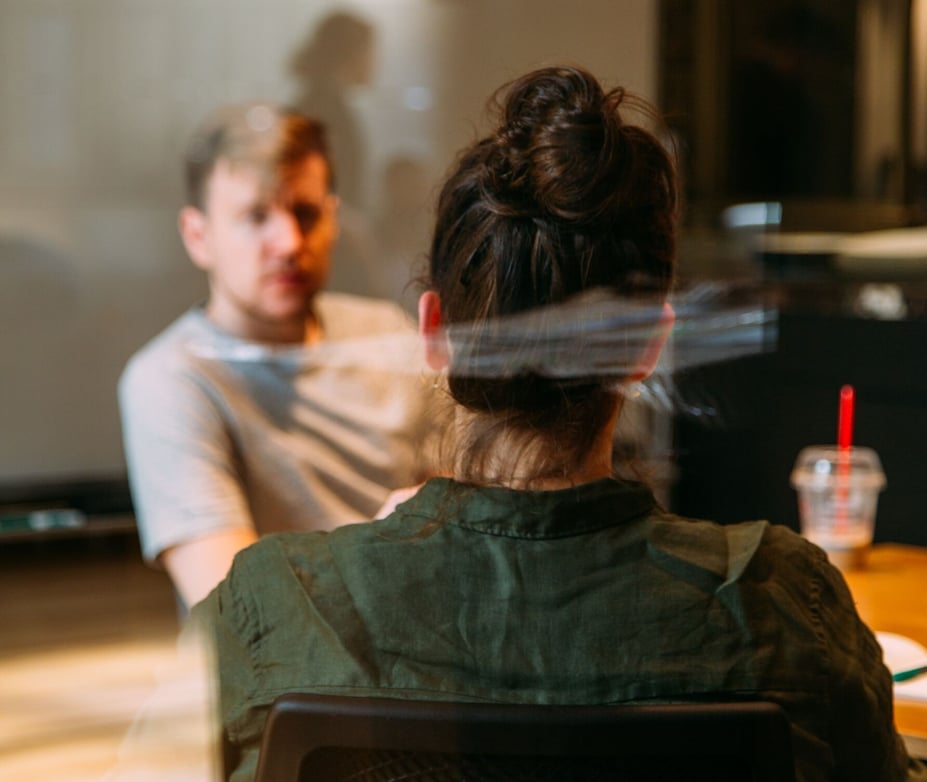 Man en vrouw zijn in gesprek. We kijken over de schouder van de vrouw mee en zien haar gesprekspartner, die niet in focus is.