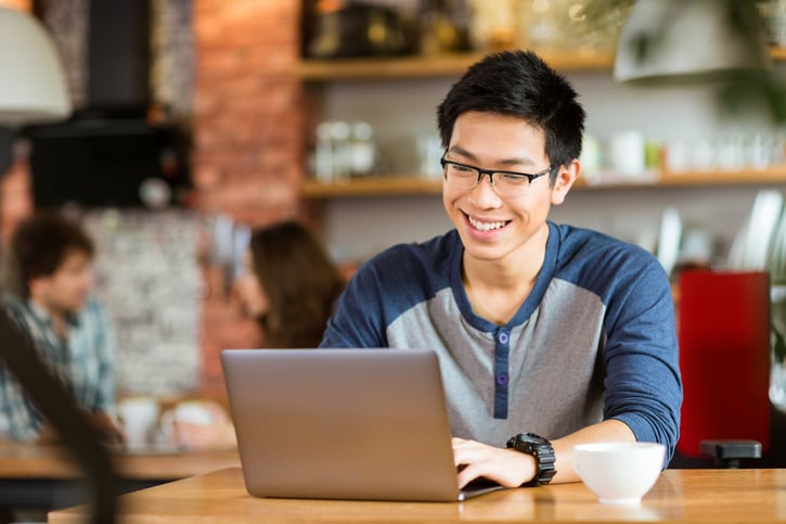 Man zit in een koffie cafe, draagt een bril en glimlacht terwijl hij naar zijn laptop scherm kijkt