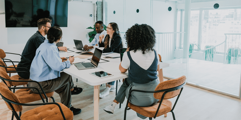 In der Bildaufnahme ist ein Meeting zu sehen, das während der Kernarbeitszeit in einem modernen Arbeitsplatz stattfindet. Mehrere Mitarbeiter sitzen um einen Tisch und diskutieren lebhaft, während sie ihre Arbeitszeitregelungen und die Bedeutung der Anwesenheitspflicht im Unternehmen erörtern.