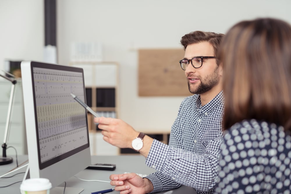 Empleado con anteojos y camisa a cuadros, explicando un análisis de negocios mostrado en el monitor de una PC de escritorio a su colega