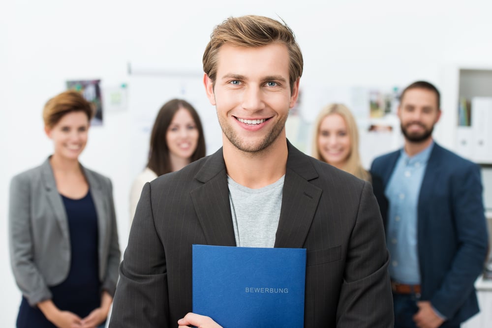 Successful smiling young male job applicant holding a blue file with his curriculum vitae posing in front of his new work colleagues or business team-3