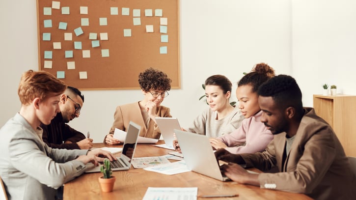 Im Bild ist eine Gruppe von Mitarbeitenden und HR-Mitarbeitern zu sehen, die im Büro an der Dienstplanung im Team-Rhythmus arbeiten. Sie diskutieren über Daten und Rollen, während im Hintergrund ein Warteraum mit Informationen zu Veranstaltungen und Tickets zu sehen ist.