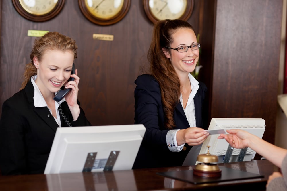 Two beautiful young stylish receptionists at a reception desk, one talking on the telephone and the other handing a card to a customer-1