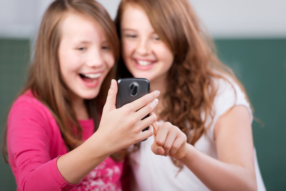 Erste Generation Y sowie  Generation Alpha Teenager in ihren Kinderschuhen, sitzend und gemeinsam konzentriert auf ein Smartphone blickend, während sie eine neue App entdecken. Auf dem Bildschirm sind bunte Icons zu sehen, die an das Alphabet erinnern.