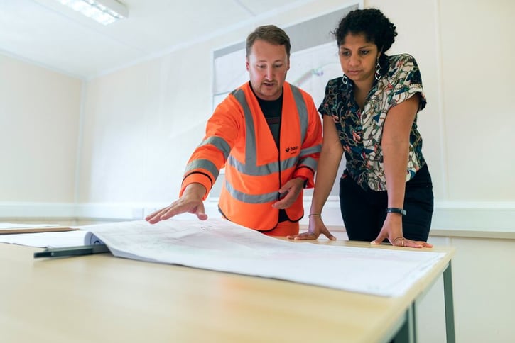 Man in orange engineering vest and woman are standing at a table and deliberating.