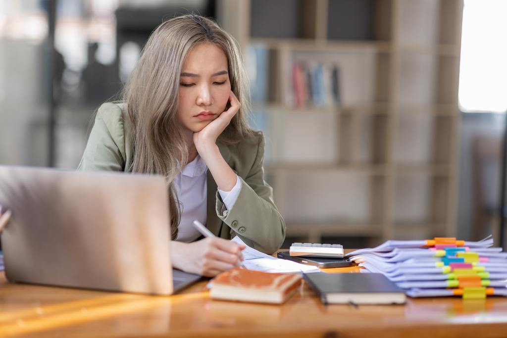 bored young business asian woman sitting at her work desk with stacks of paper on her desk