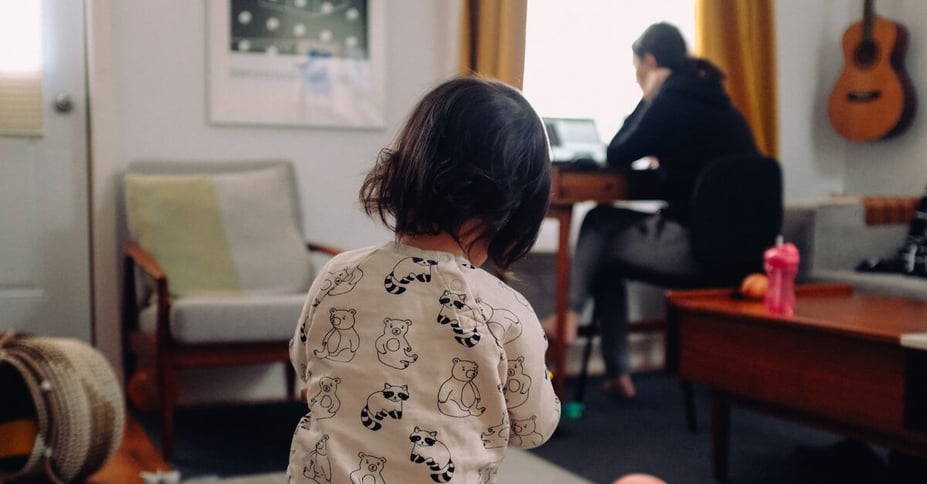Kindje staat met rug naar camera toegekeerd terwijl moeder aan bureau zit te werken op de achtergrond