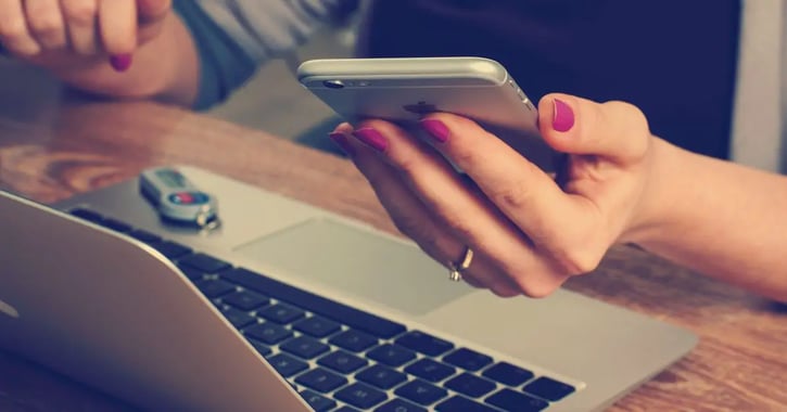 Vrouw zit voor laptop en kijkt tegelijkertijd op haar telefoon