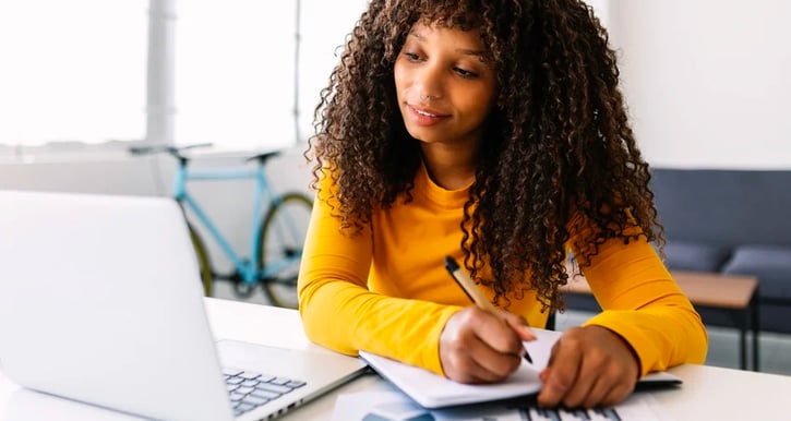 Vrouw zit voor laptop, schrijft op kladblok naast laptop. Er staat een blauwe fiets tegen de muur op de achtergrond