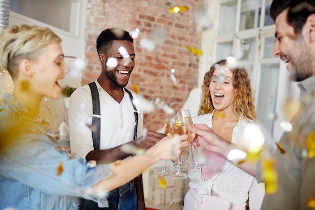Gratulation: Gruppe von Kollegen die in einem festlich dekorierten Büro zusammenstehen und mit Sekt auf die Beförderung eines Teammitglieds anstoßen während der Beförderte eine Glückwunschkarte und ein Geschenk in den Händen hält