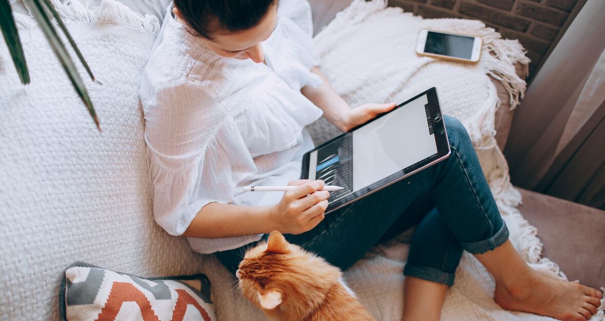 Vrouw zit op een bank en gebruikt een tablet terwijl de kat toekijkt