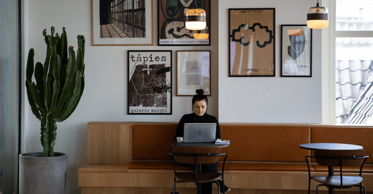 Vrouw zit op een bank en aan een tafeltje met haar laptop en een kop koffie voor zich.