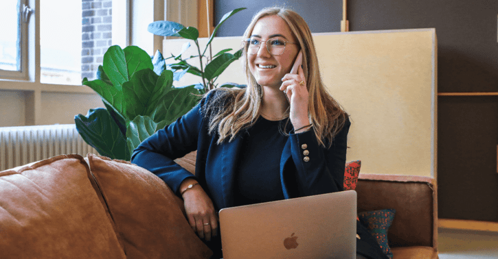 Vrouw zit op de bank met een laptop op haar schoot. Ze is aan de telefoon en ze lacht,