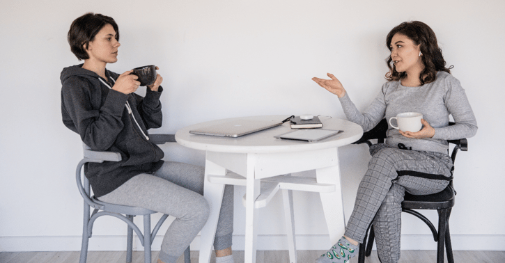 Twee vrouwen zitten aan tafel met een kopje in hun handen en praten.