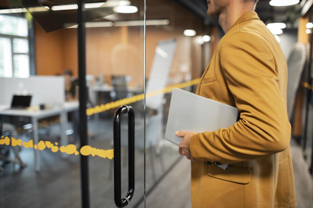 man wearing a mustard colored blazer walking into meeting room while holding laptop 
