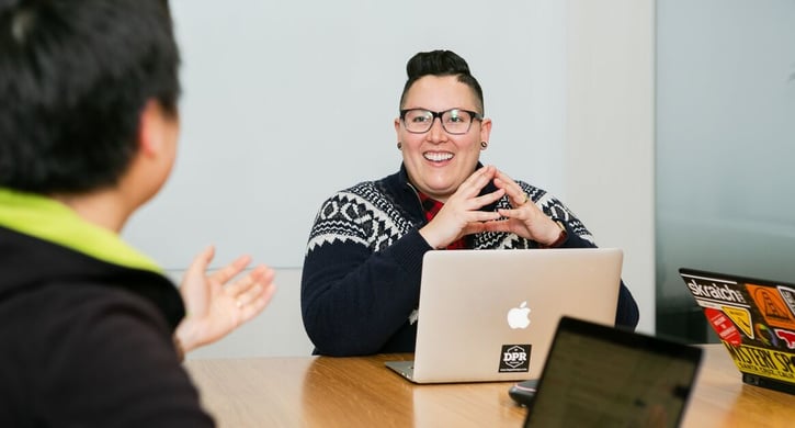 Persoon zit aan tafel voor laptop en lacht naar collega.
