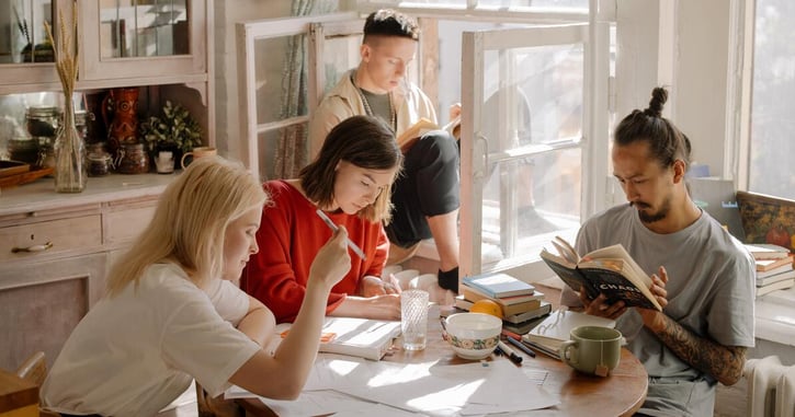 Mensen zitten samen aan een tafel te leren en te lezen. 1 iemand zit op de achtergrond in de vensterbank 