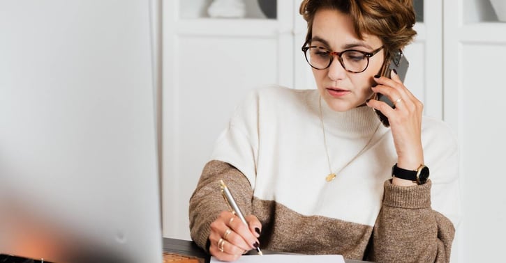 Vrouw zit aan bureau met een telefoon aan haar oor en schrijft