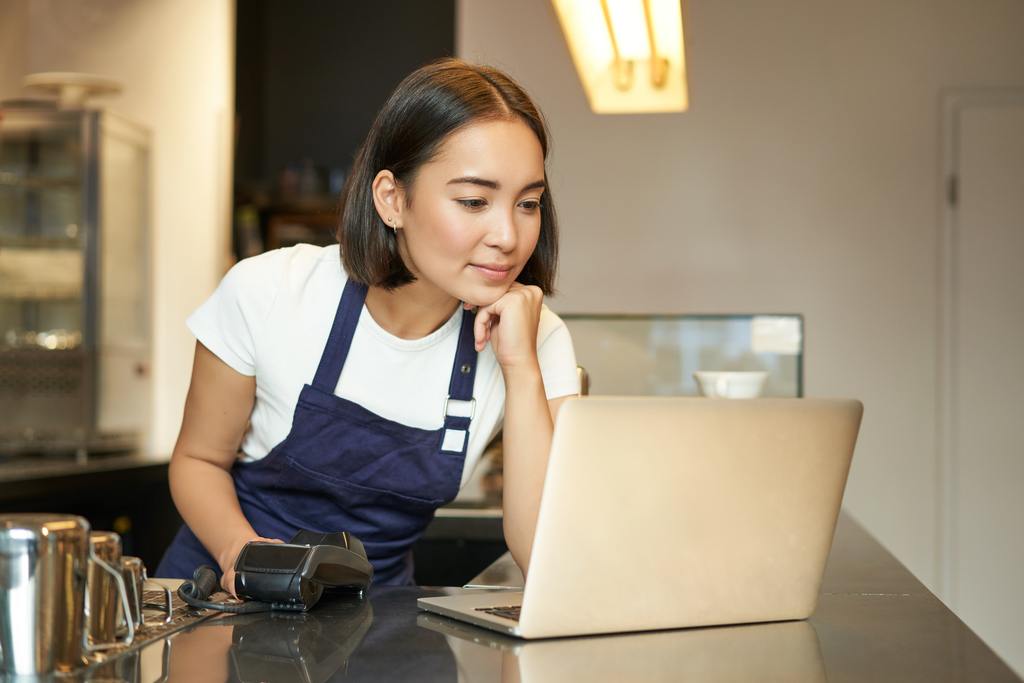 smiling asian female bar manager or employee in cafe looking at laptop