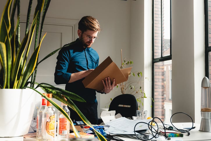 Man met bril staat achter een rommelig bureau naar een document in zijn handen te kijken