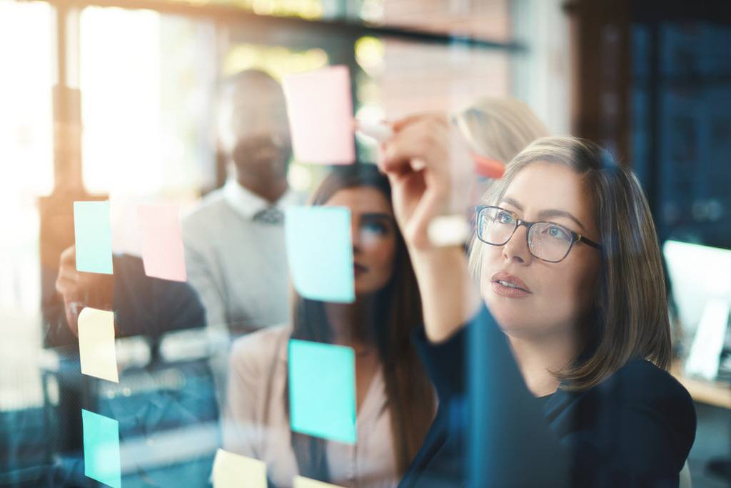 coworkers writing on sticky note on a glass board during team meeting